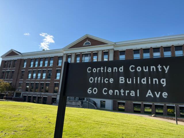 Brick office building with white trim and arched window, under a blue sky; foreground sign reads Cortland County Office Building, 60 Central Ave (partial).