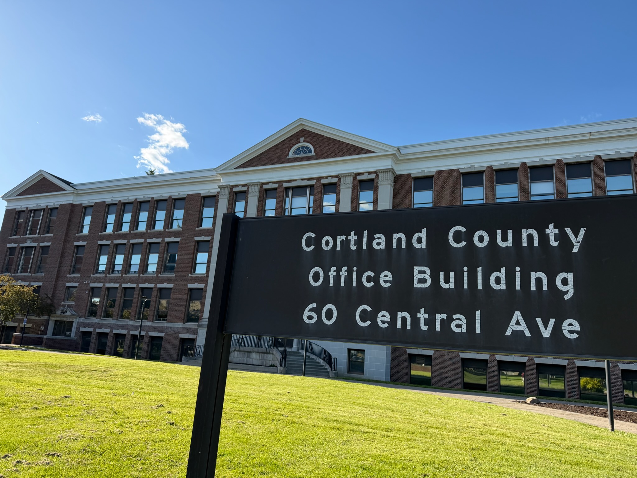 Brick office building with white trim and arched window, under a blue sky; foreground sign reads Cortland County Office Building, 60 Central Ave (partial).