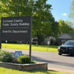 Large sign reads 'Cortland County Public Safety Building' with 'Sheriffs Department' beneath; a sheriff SUV parked nearby on a sunny day.