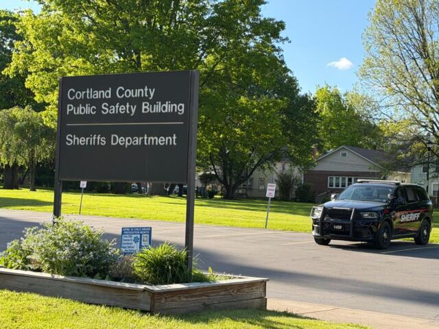 Large sign reads 'Cortland County Public Safety Building' with 'Sheriffs Department' beneath; a sheriff SUV parked nearby on a sunny day.