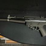 Long-barreled rifle with a suppressor resting on a dark surface, left side visible.