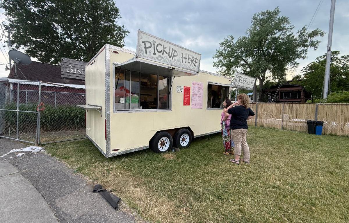 Food truck trailer with a large 'PICKUP HERE' sign, serving customers at a grassy lot.