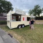 Food truck trailer with a large 'PICKUP HERE' sign, serving customers at a grassy lot.