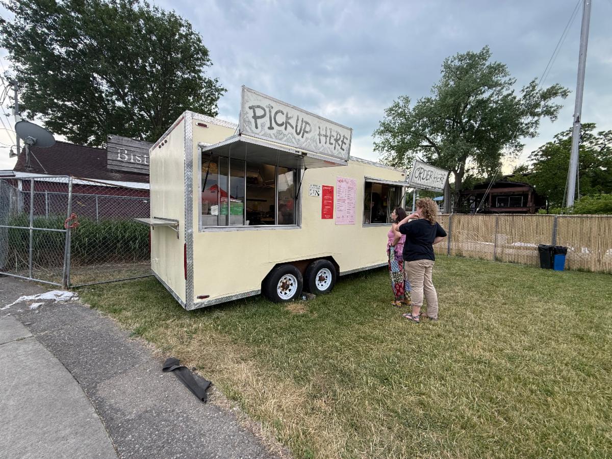 Food truck trailer with a large 'PICKUP HERE' sign, serving customers at a grassy lot.