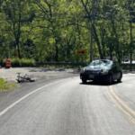 Black SUV navigating a sharp curve on a forested rural road, with orange construction barrels and a toppled trailer on the left near barricades.