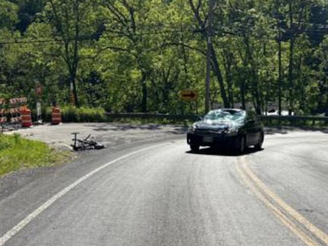 Black SUV navigating a sharp curve on a forested rural road, with orange construction barrels and a toppled trailer on the left near barricades.