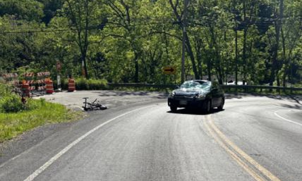 Black SUV navigating a sharp curve on a forested rural road, with orange construction barrels and a toppled trailer on the left near barricades.