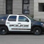 Police SUV parked on a city street in front of a stone government building; POLICE and City of Elmira markings on the side.