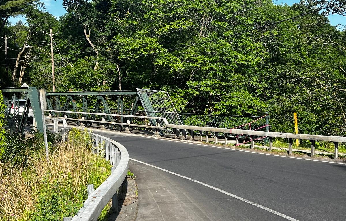 Curved asphalt road crossing a green metal truss bridge, with guardrails and dense trees beyond.