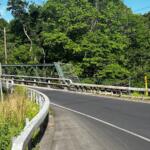 Curved asphalt road crossing a green metal truss bridge, with guardrails and dense trees beyond.