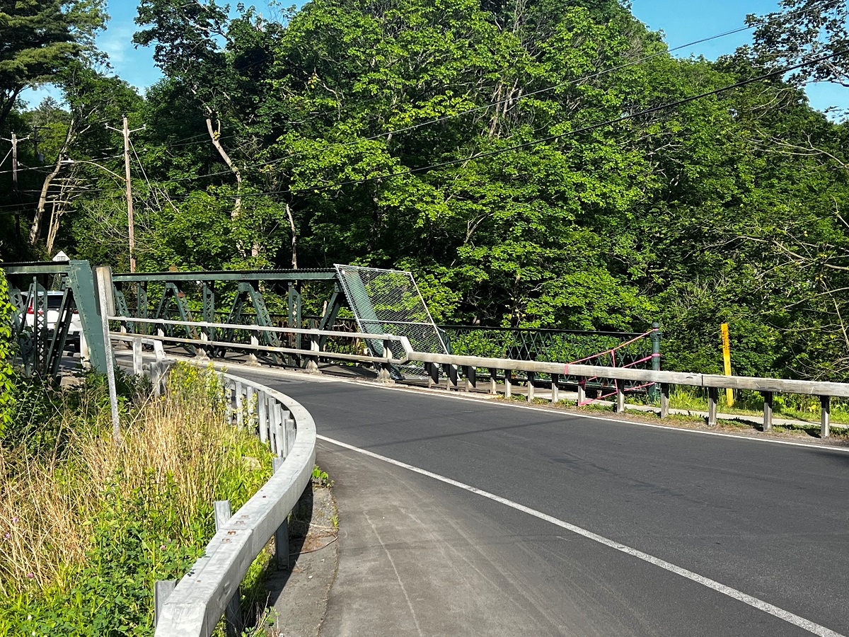 Curved asphalt road crossing a green metal truss bridge, with guardrails and dense trees beyond.