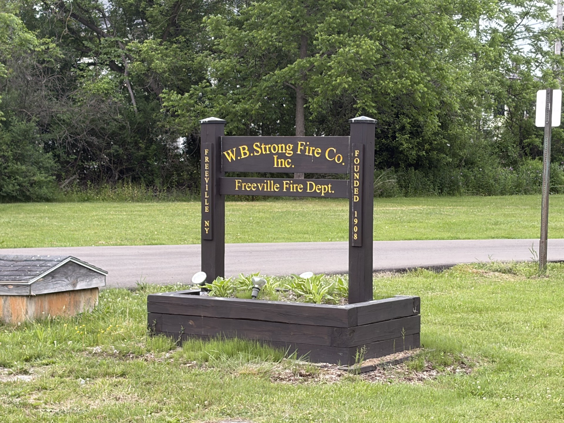 Wooden sign for W.B. Strong Fire Co. Inc. and Freeville Fire Dept. with gold lettering, set in a grassy area with trees behind.