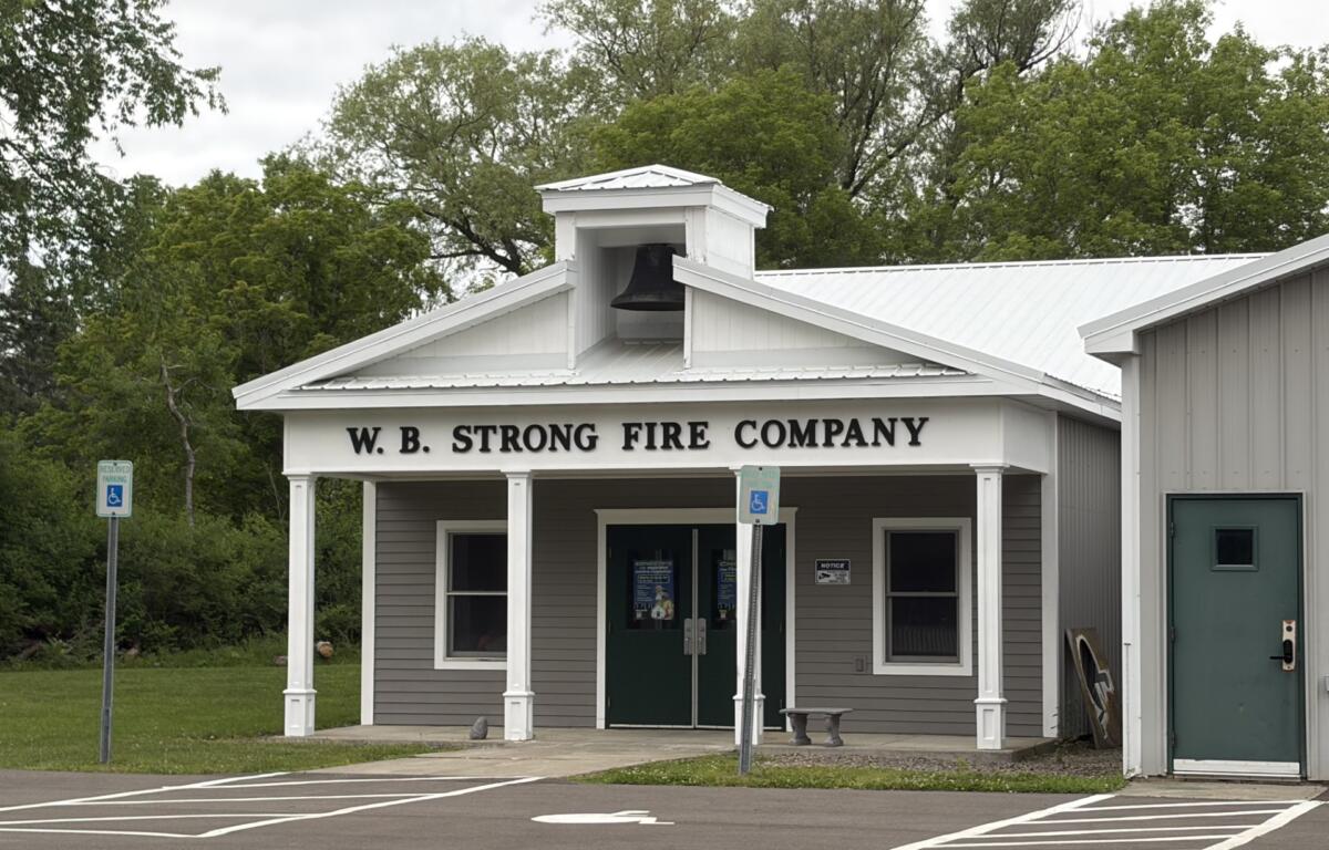 Fire station with a white cupola bell tower and 'W.B. STRONG FIRE COMPANY' on the façade, surrounded by trees and a parking lot with wheelchair signs.