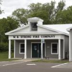 Fire station with a white cupola bell tower and 'W.B. STRONG FIRE COMPANY' on the façade, surrounded by trees and a parking lot with wheelchair signs.