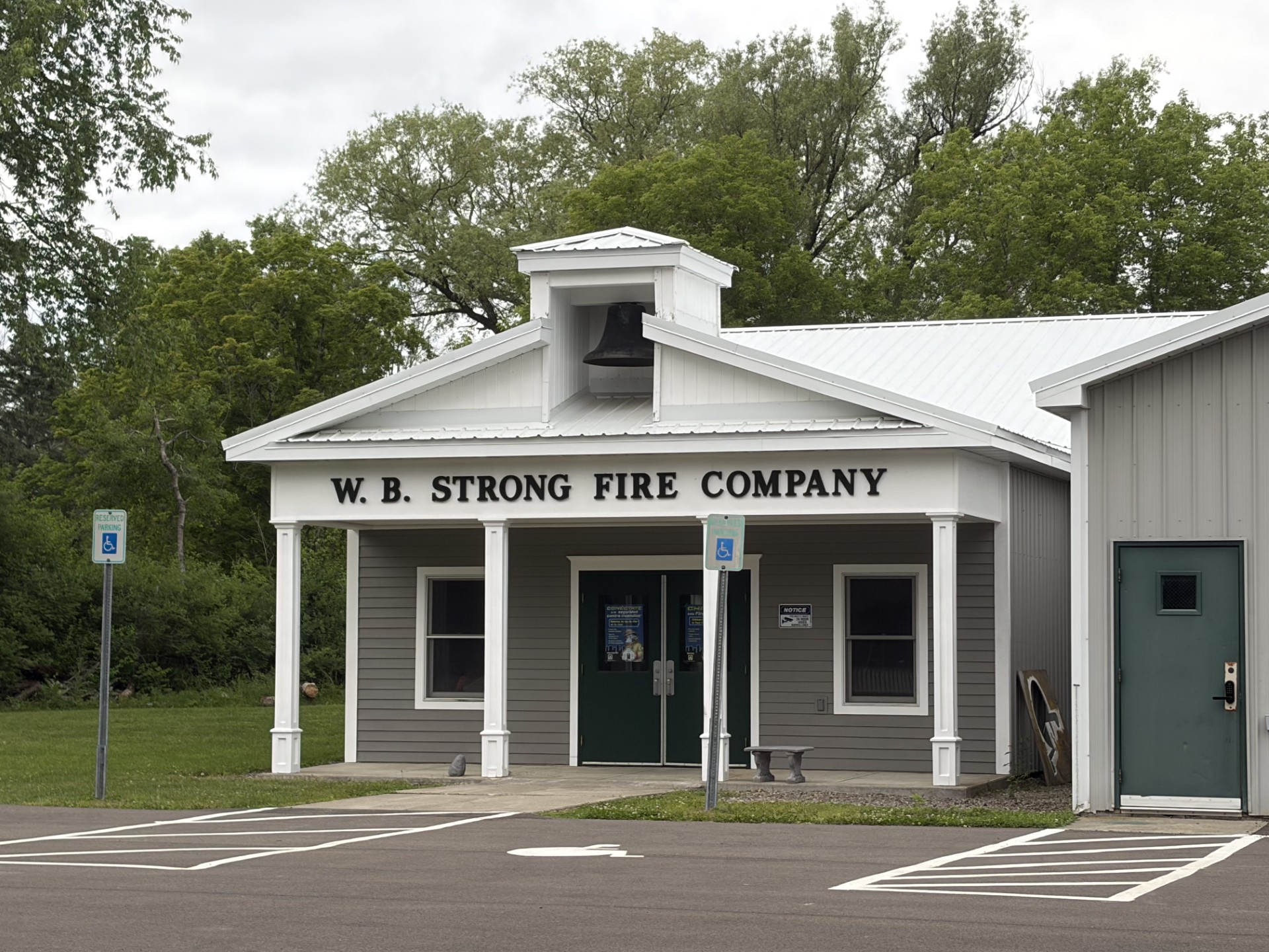Fire station with a white cupola bell tower and 'W.B. STRONG FIRE COMPANY' on the façade, surrounded by trees and a parking lot with wheelchair signs.