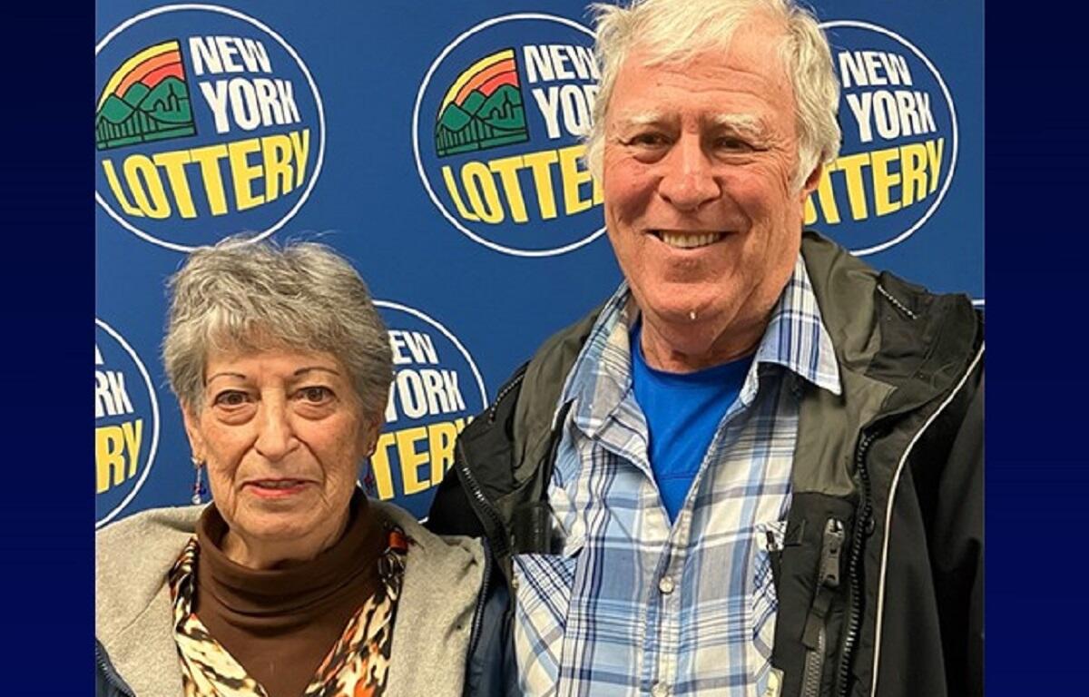 Older couple posing for a photo at a New York Lottery event with a blue backdrop and logos nearby.