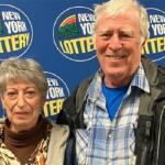 Older couple posing for a photo at a New York Lottery event with a blue backdrop and logos nearby.