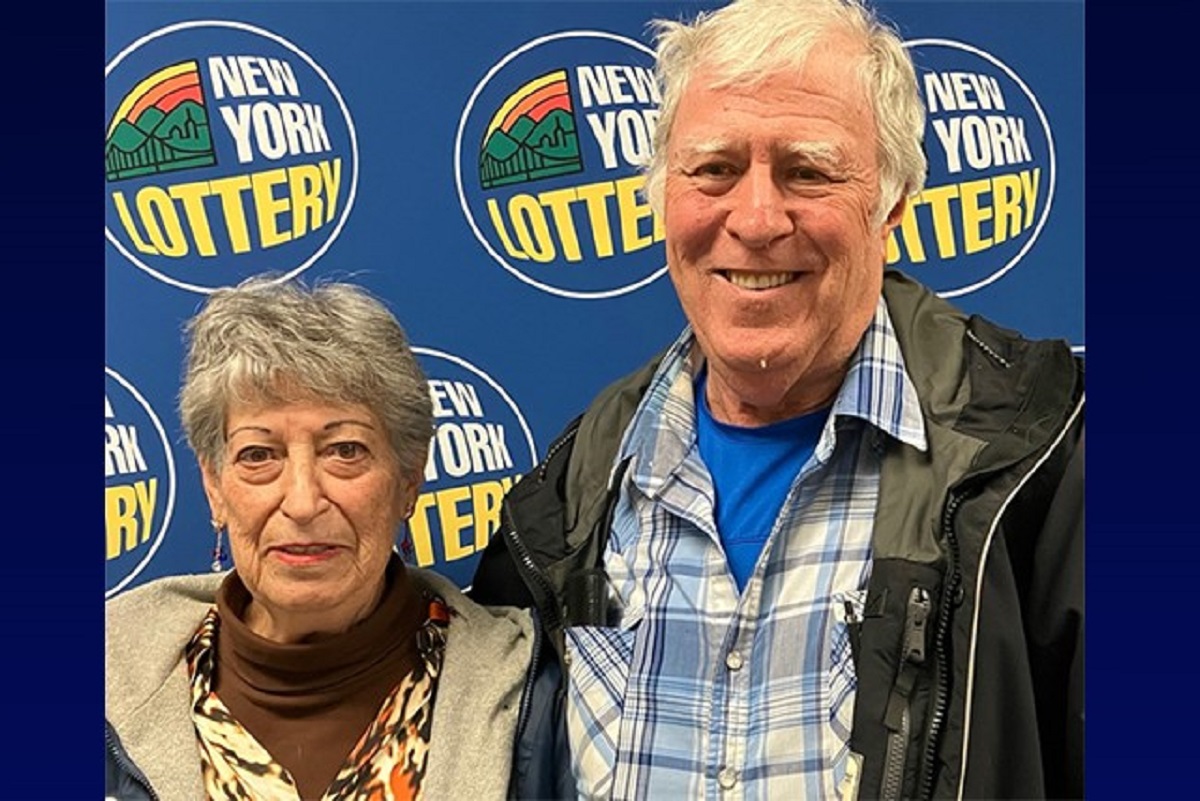 Older couple posing for a photo at a New York Lottery event with a blue backdrop and logos nearby.