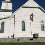 White wooden church facade with steep gables, tall arched windows, and a red abstract wall sign against a clear blue sky.