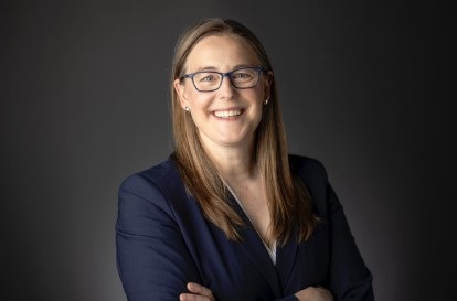 Professional headshot of a smiling woman with glasses wearing a navy blazer, arms crossed in front of a dark background.