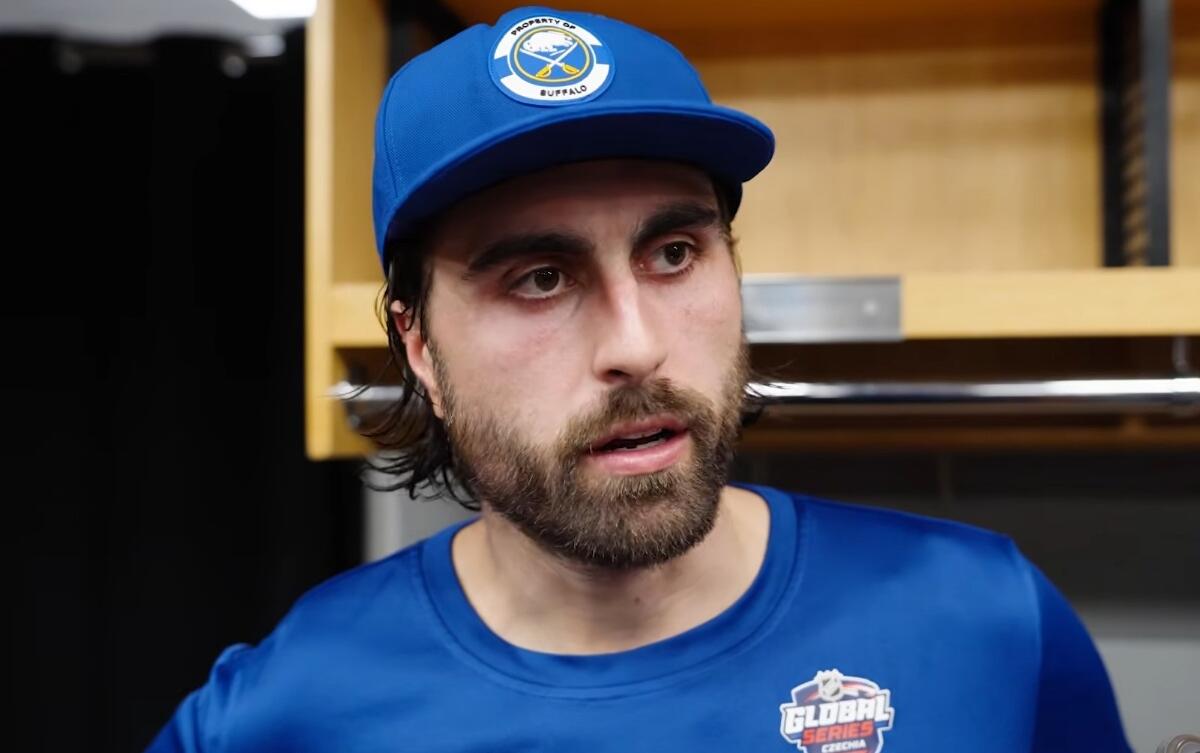Man with beard wearing a blue cap and matching shirt in a locker room, looking to the side.