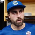Man with beard wearing a blue cap and matching shirt in a locker room, looking to the side.