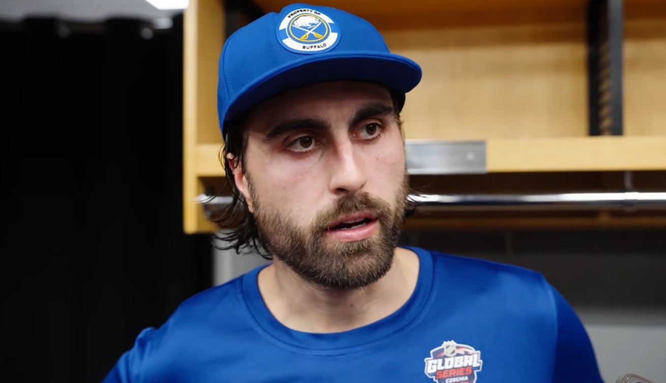 Man with beard wearing a blue cap and matching shirt in a locker room, looking to the side.