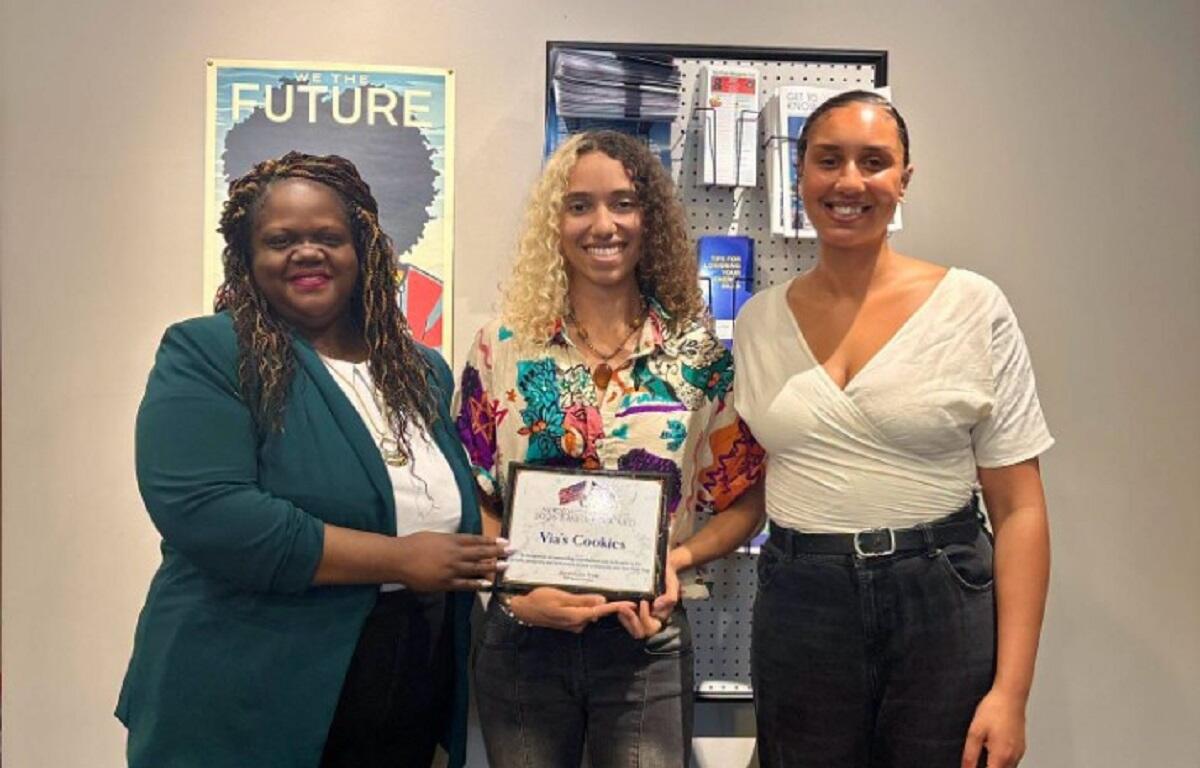 Three women stand together indoors; the center woman holds a framed award reading 'Via's Cookies' while her colleagues smile nearby.