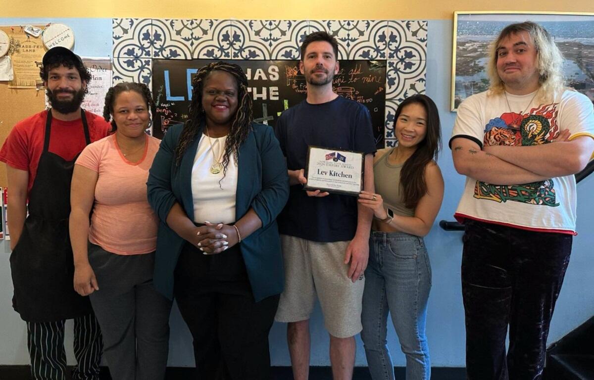 Six people posing together in a room, one man holding a framed award plaque that reads 'Lev Kitchen,' everyone smiling at the camera with a blue patterned wall behind them.