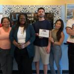 Six people posing together in a room, one man holding a framed award plaque that reads 'Lev Kitchen,' everyone smiling at the camera with a blue patterned wall behind them.