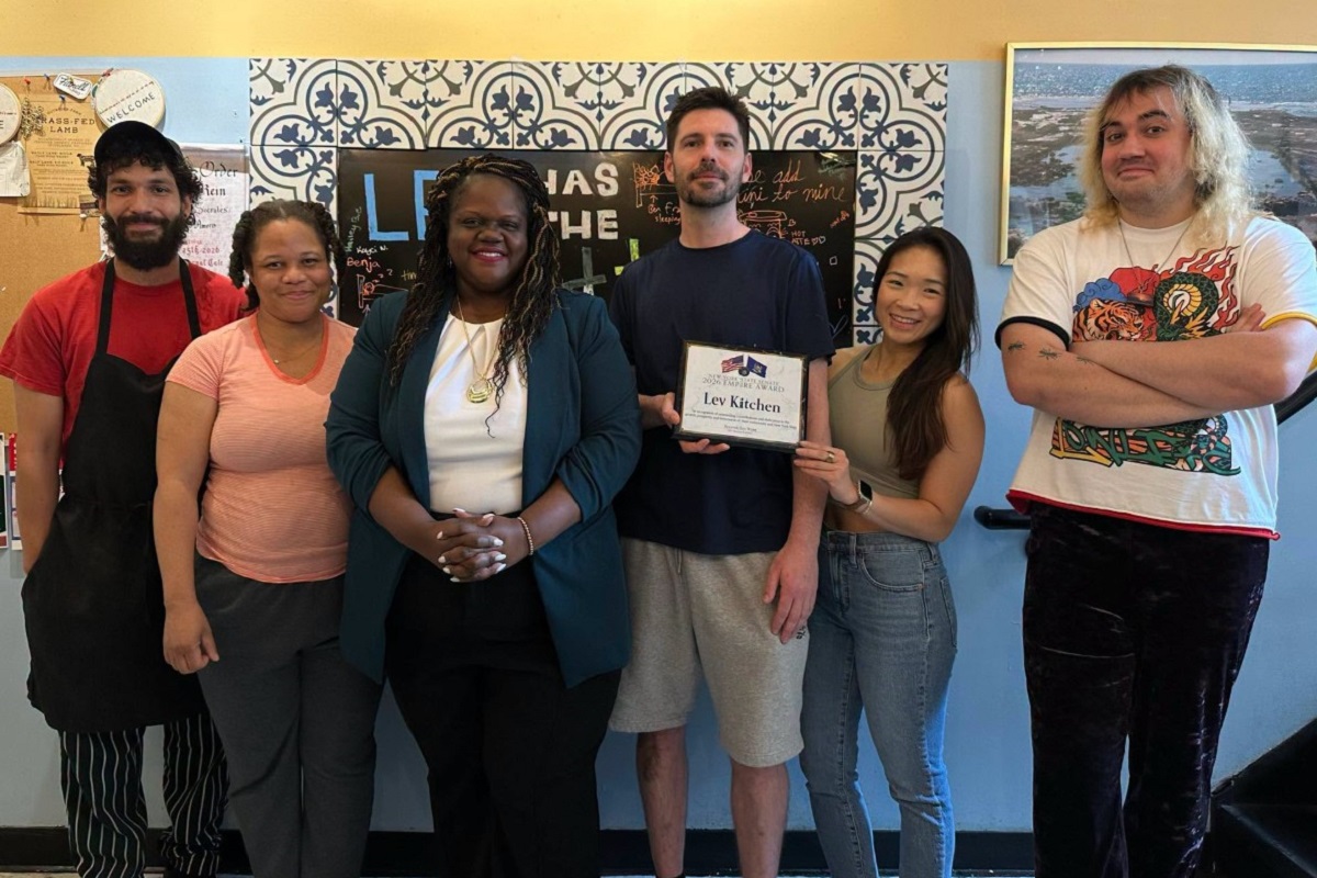 Six people posing together in a room, one man holding a framed award plaque that reads 'Lev Kitchen,' everyone smiling at the camera with a blue patterned wall behind them.