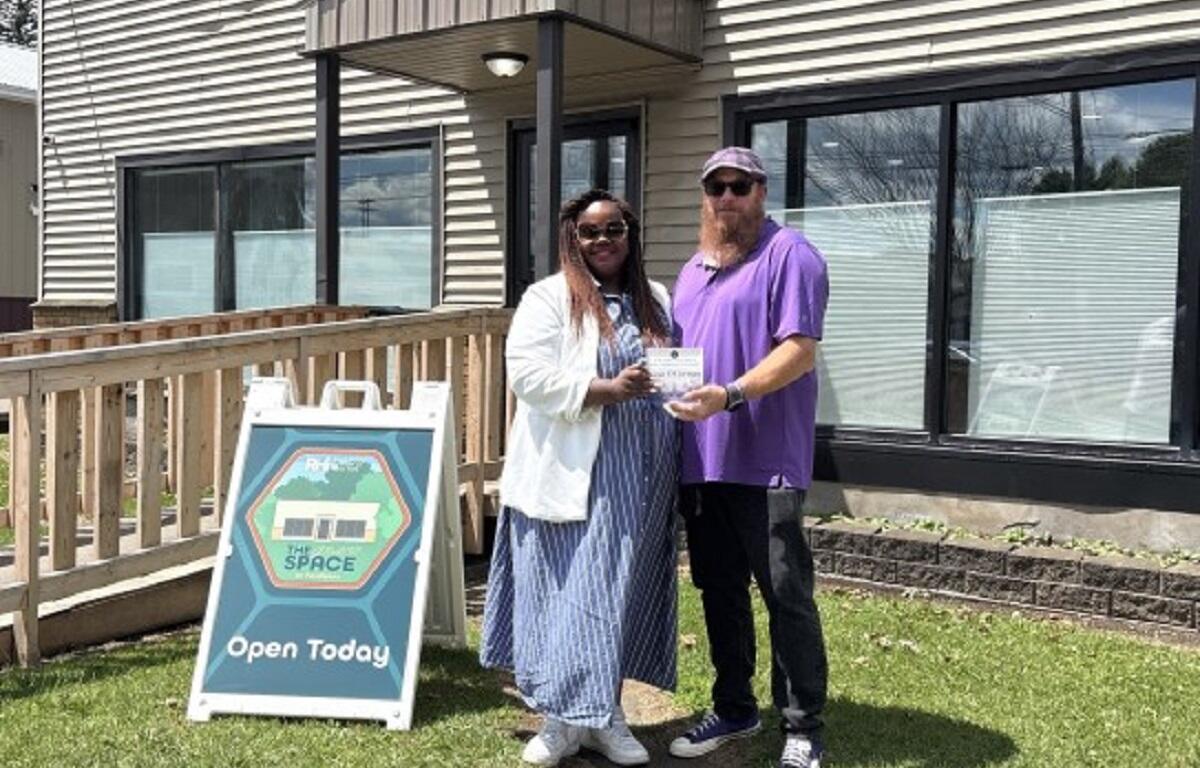 Two people pose outside a building as one hands a certificate to the other, with an 'Open Today' sign nearby in the yard.