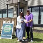 Two people pose outside a building as one hands a certificate to the other, with an 'Open Today' sign nearby in the yard.