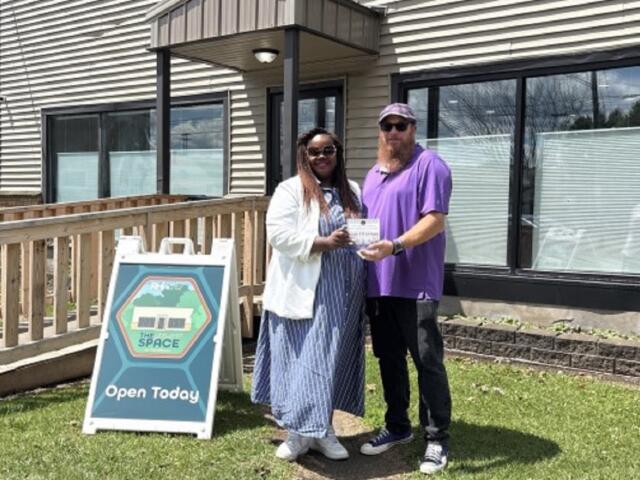 Two people pose outside a building as one hands a certificate to the other, with an 'Open Today' sign nearby in the yard.