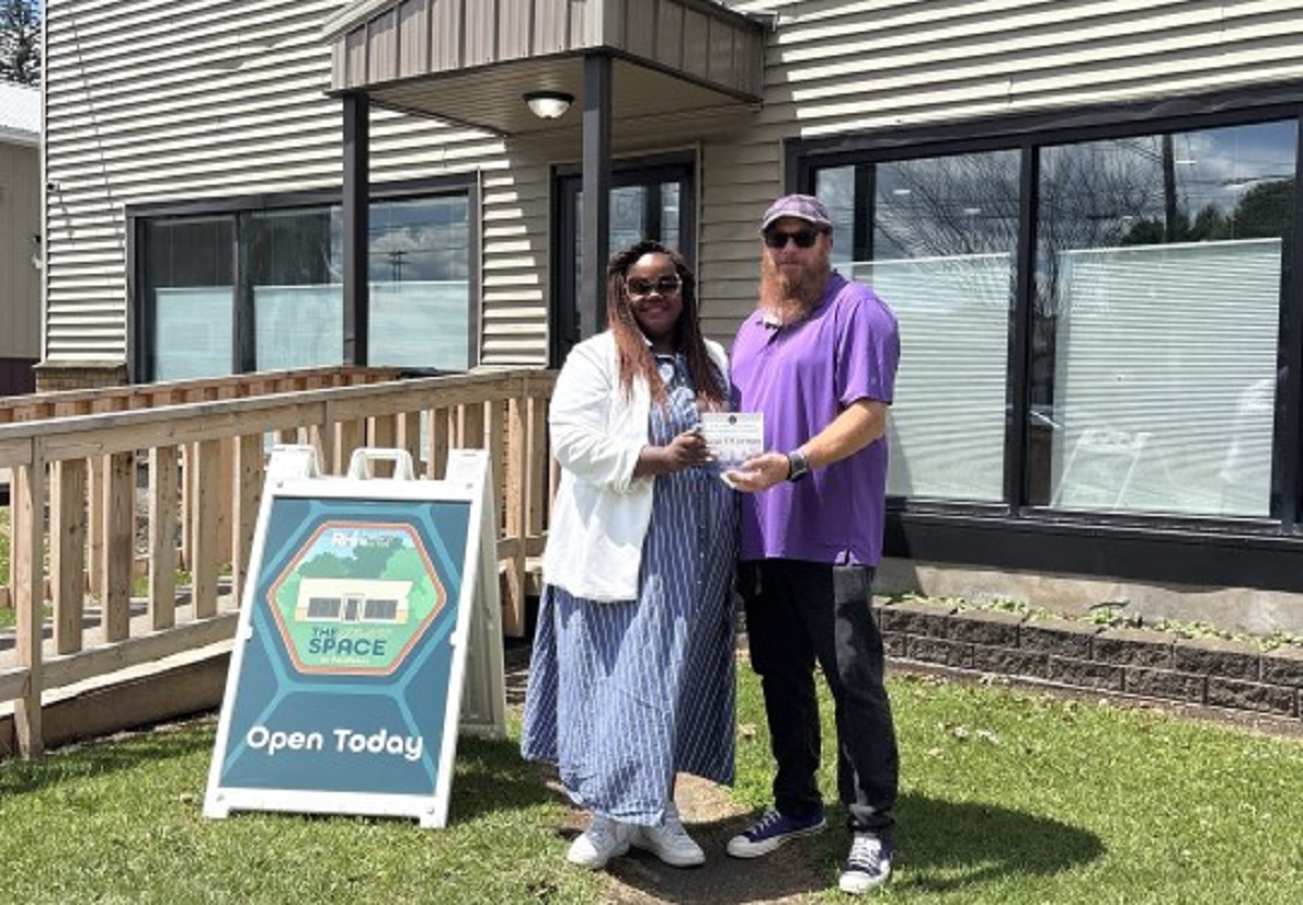 Two people pose outside a building as one hands a certificate to the other, with an 'Open Today' sign nearby in the yard.