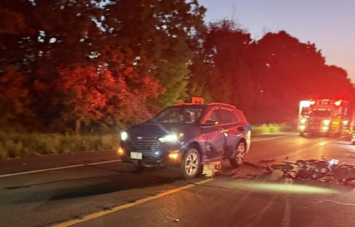 Blue SUV with front/side damage sits in the road after a crash, with motorcycle debris scattered in front and a red-emergency vehicle glowing in the distance at dusk.