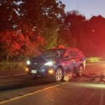 Blue SUV with front/side damage sits in the road after a crash, with motorcycle debris scattered in front and a red-emergency vehicle glowing in the distance at dusk.