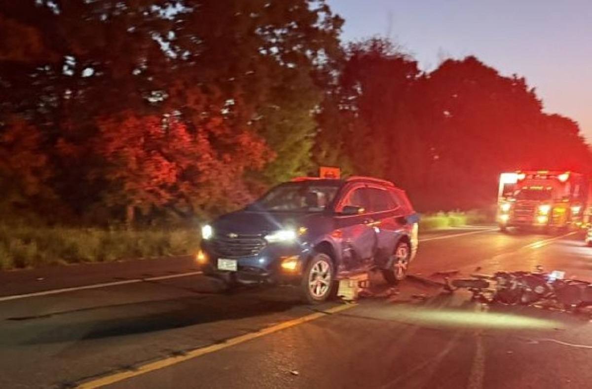 Blue SUV with front/side damage sits in the road after a crash, with motorcycle debris scattered in front and a red-emergency vehicle glowing in the distance at dusk.
