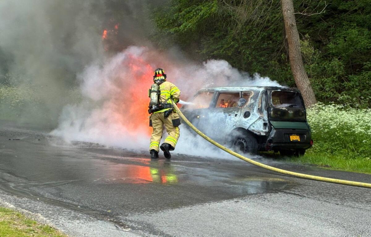 Firefighter in turnout gear hoses a burning van on a roadside, thick smoke and flames rising behind.