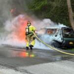 Firefighter in turnout gear hoses a burning van on a roadside, thick smoke and flames rising behind.