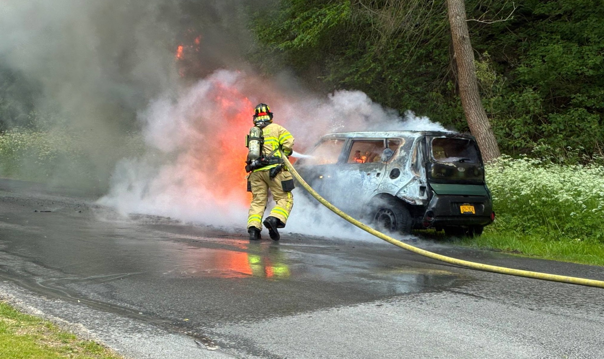 Firefighter in turnout gear hoses a burning van on a roadside, thick smoke and flames rising behind.