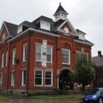 Red brick town-style building with white trim, arched windows, and a small cupola on a rainy street.