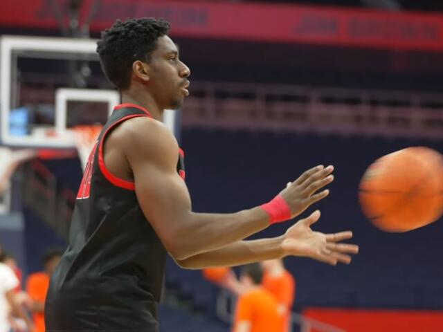 Basketball player in a black uniform passing an orange ball on a court with spectators in the background.