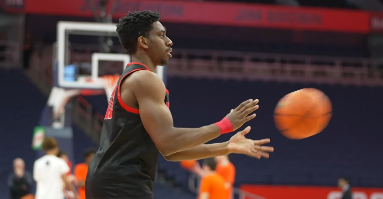 Basketball player in a black uniform passing an orange ball on a court with spectators in the background.