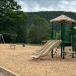 Open-air playground in a park with beige twin slides, green climbing structure, swings, and wood-chip ground near trees and hills in the background.