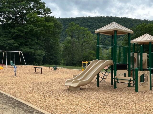 Open-air playground in a park with beige twin slides, green climbing structure, swings, and wood-chip ground near trees and hills in the background.