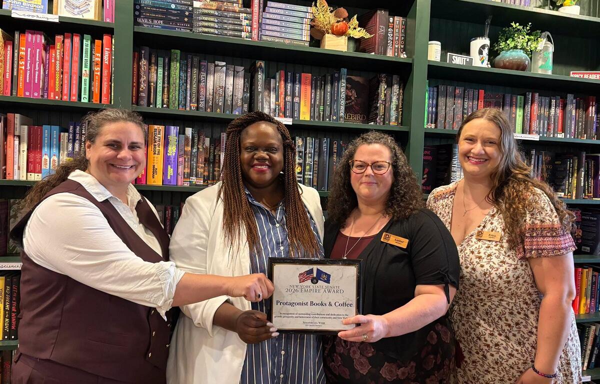 Four women in a bookstore pose with a framed award certificate, smiling, in front of tall shelves of books.