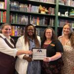 Four women in a bookstore pose with a framed award certificate, smiling, in front of tall shelves of books.