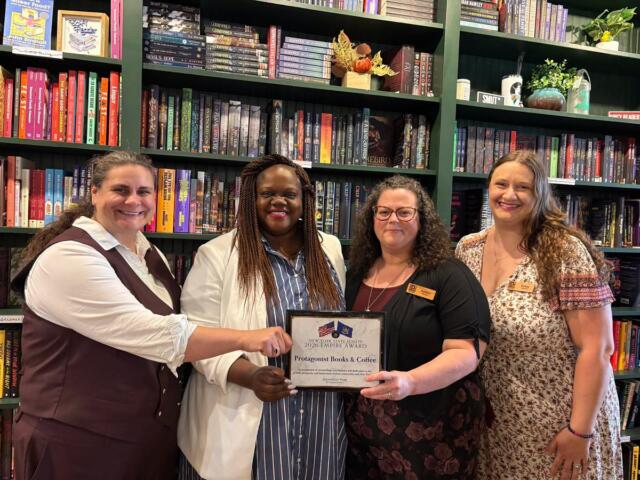 Four women in a bookstore pose with a framed award certificate, smiling, in front of tall shelves of books.