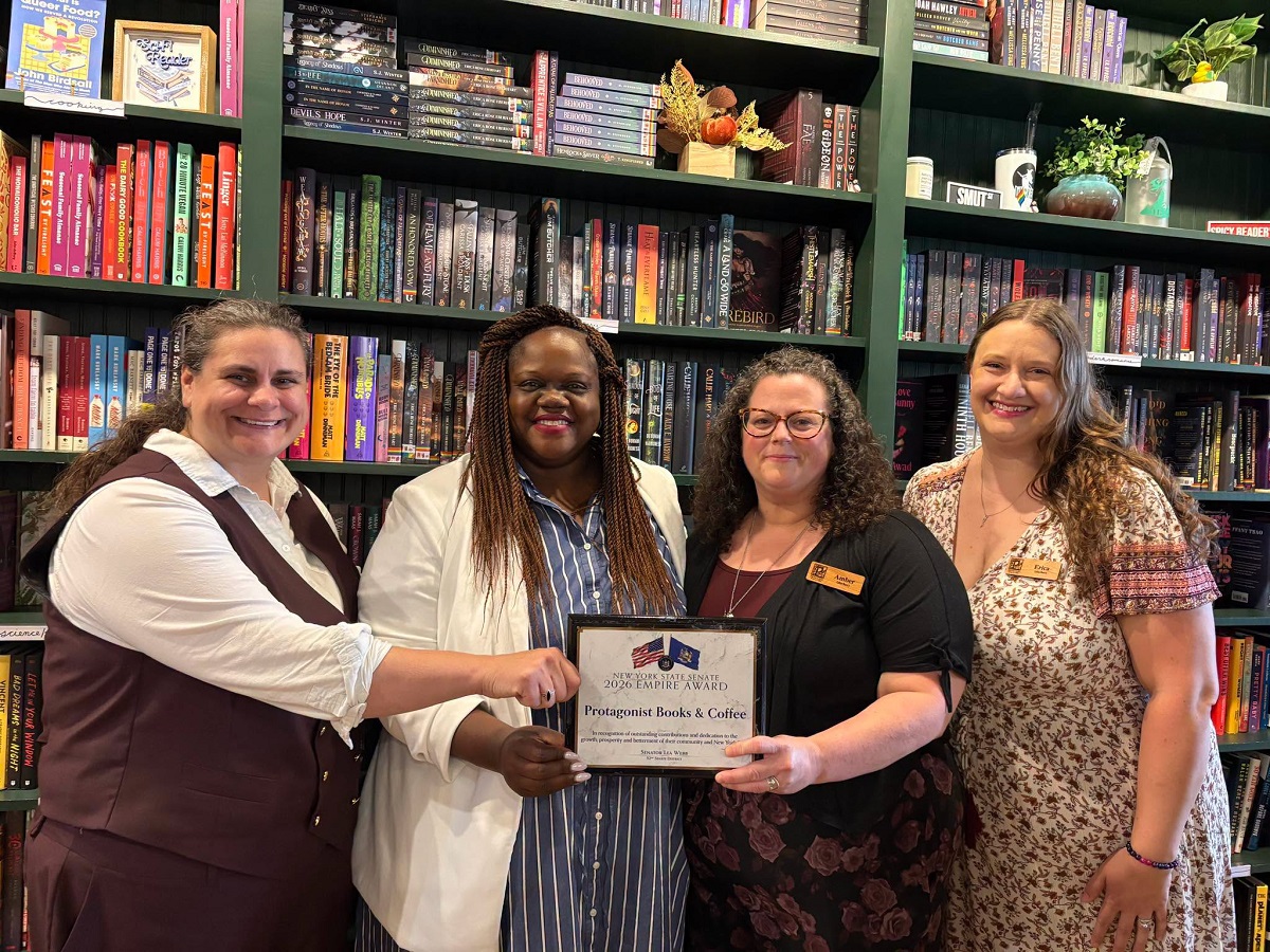 Four women in a bookstore pose with a framed award certificate, smiling, in front of tall shelves of books.